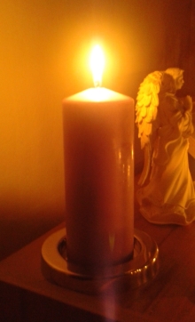 Leuchtende Stumpenkerze mit Engel im Hintergrund.